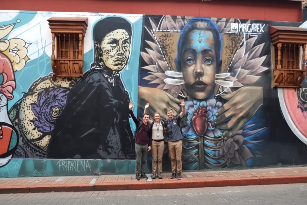 group of tourists standing in front of a vibrant street art mural featuring stylized human faces and symbolic imagery in Bogotá Colombia