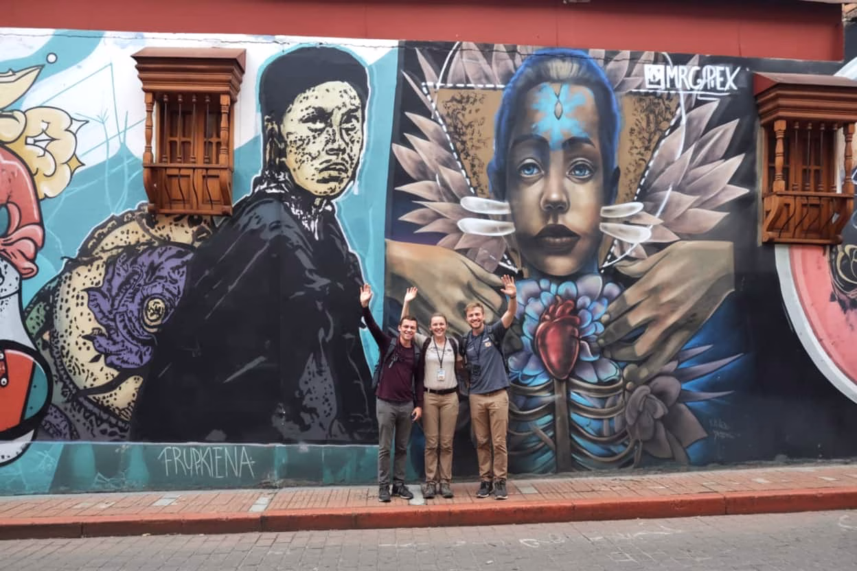 group of tourists standing in front of a vibrant street art mural featuring stylized human faces and symbolic imagery in Bogotá Colombia