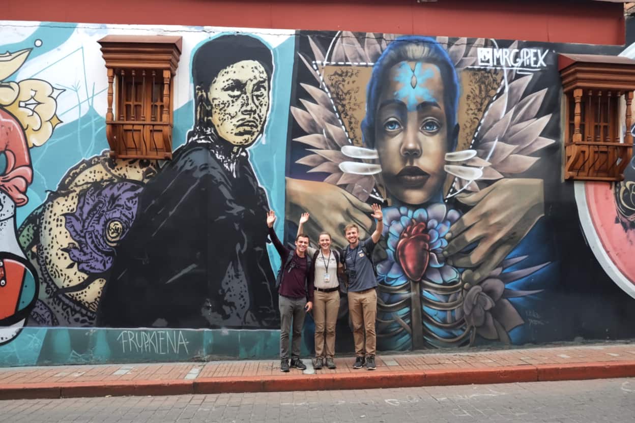 group of tourists standing in front of a vibrant street art mural featuring stylized human faces and symbolic imagery in Bogotá Colombia