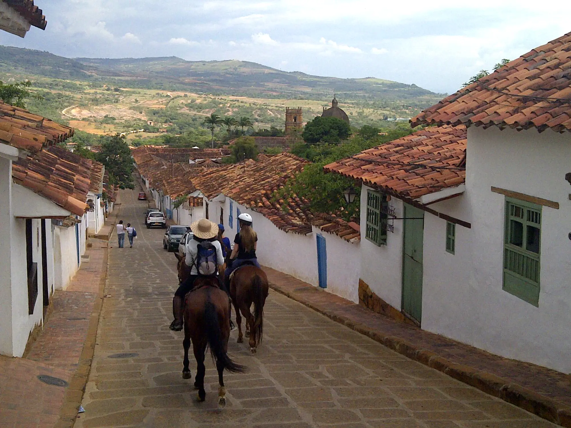 Travelers horseback riding down a cobblestone street with colonial rooftops and mountain views on a Barichara Tour.
