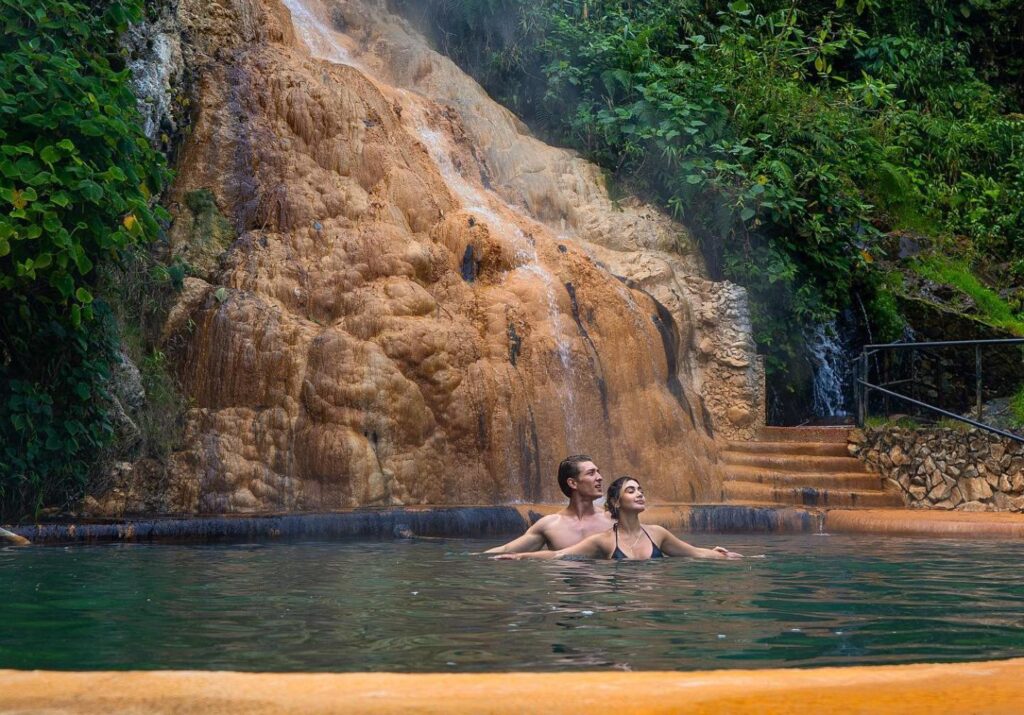 Couple relaxing in a natural thermal pool beneath a mineral waterfall at Santa Rosa de Cabal hot springs, surrounded by lush tropical forest.